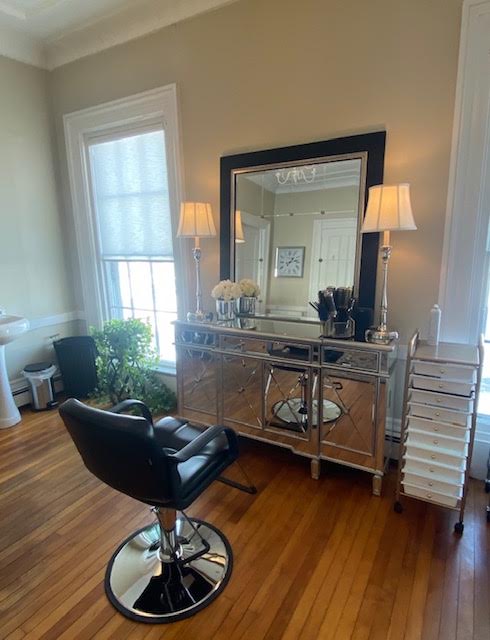 Salon styling station with a mirrored console, framed mirror, paired lamps, and a black chair on hardwood